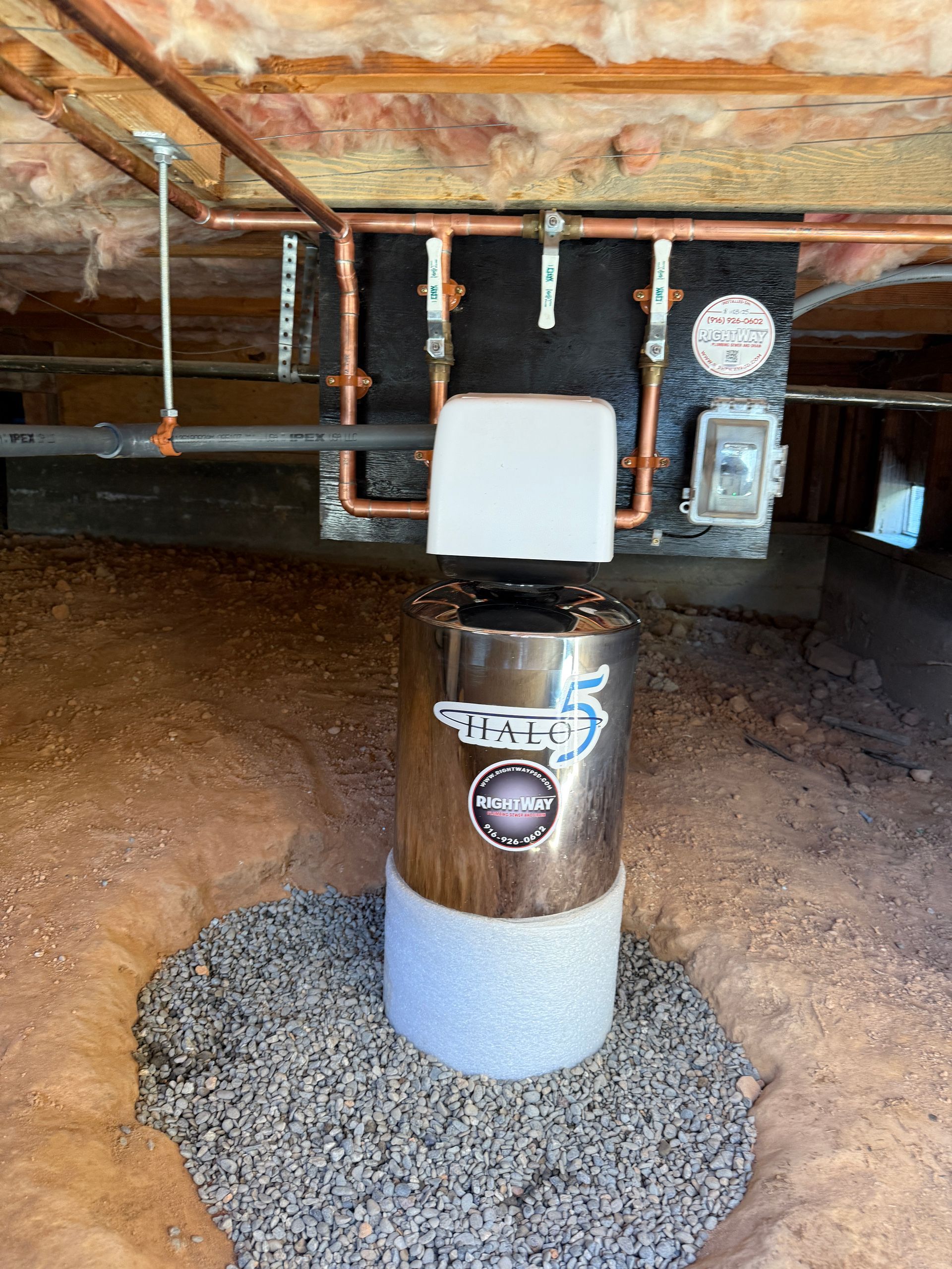 Rusty water heater and plumbing under a house, with a white box mounted on top and gravel below