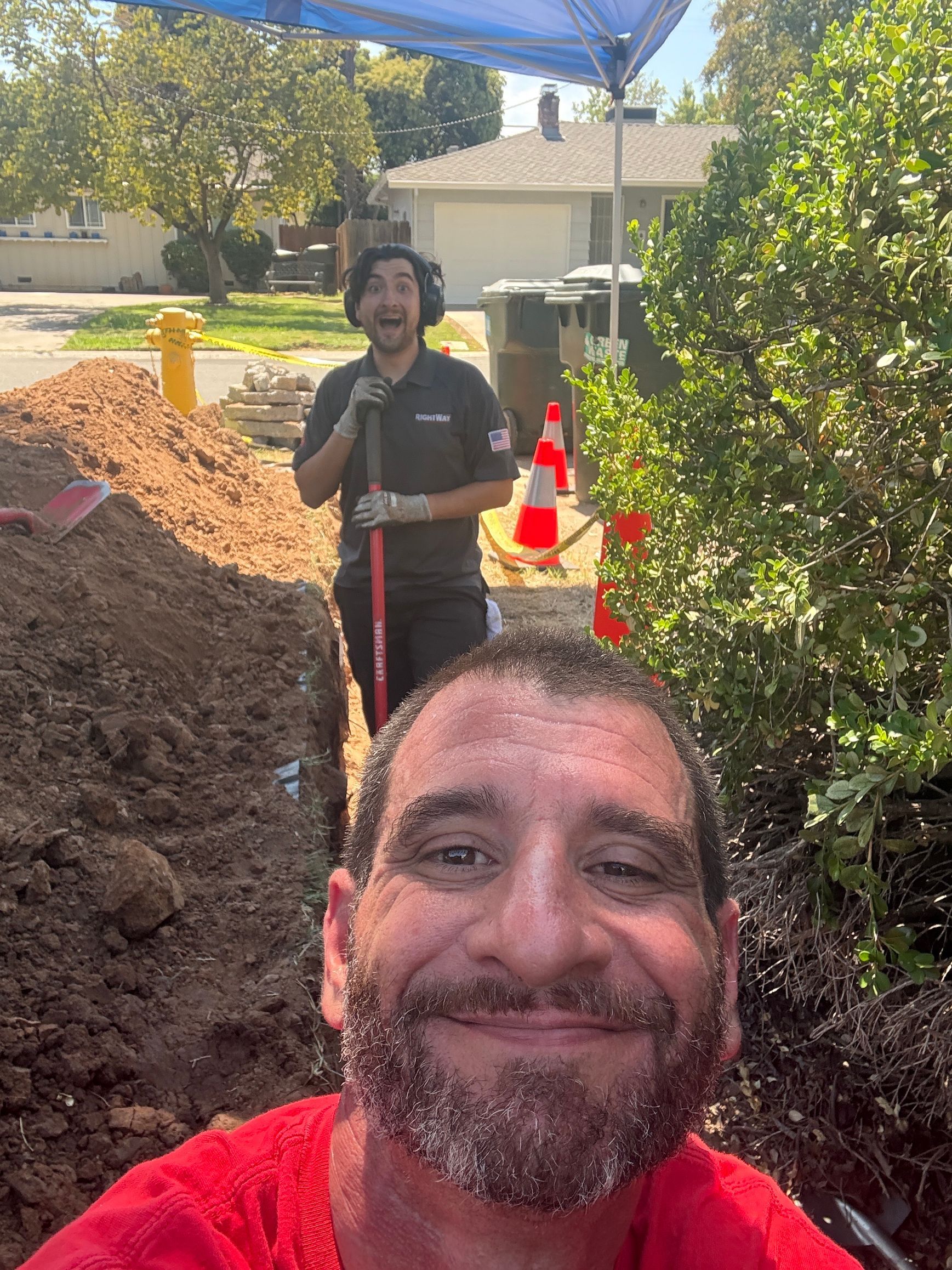 Two men working in a trench; one taking a selfie. Outdoors, dirt, safety cones, blue tent.
