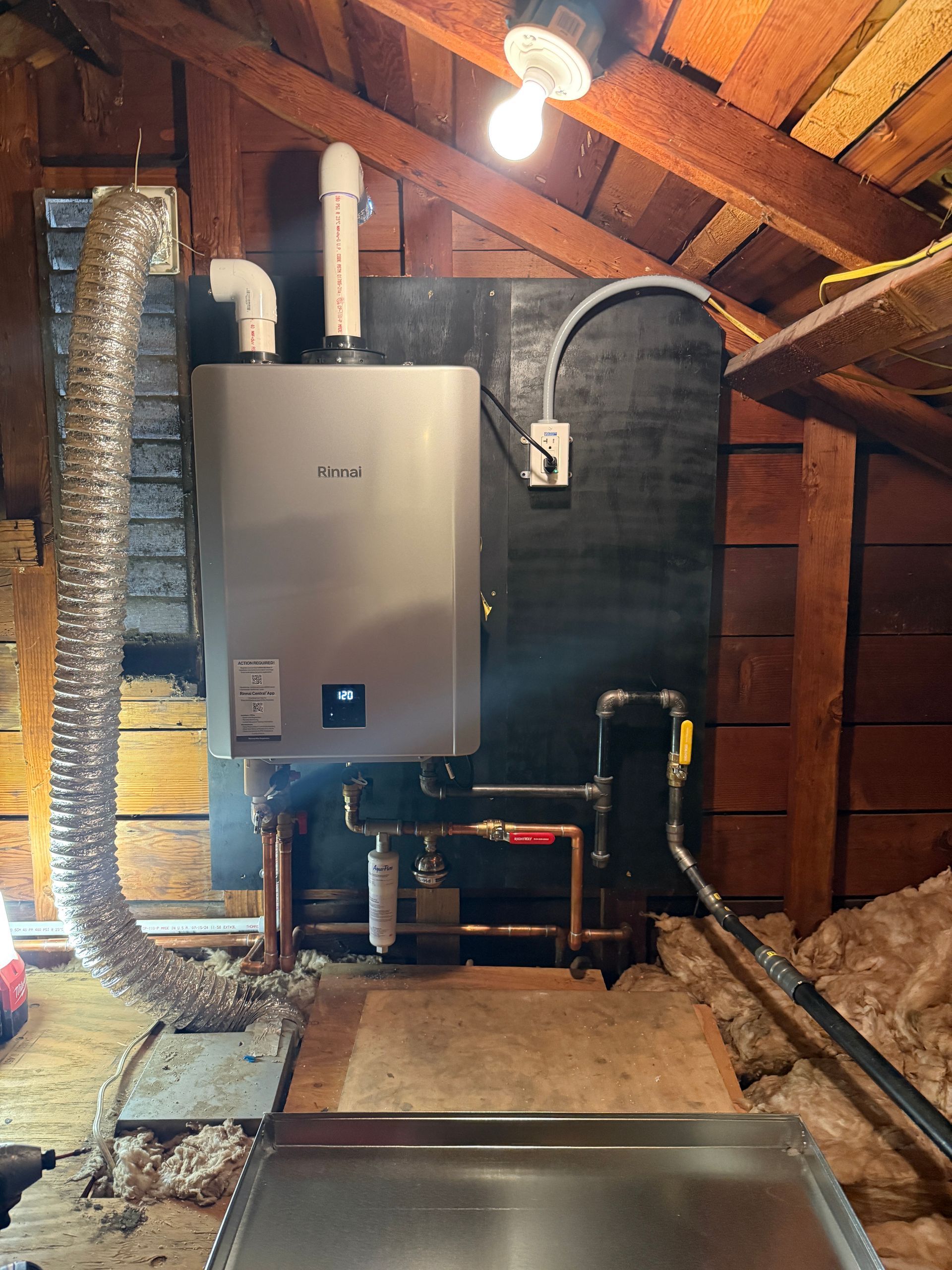 Rightway Plumbing Sewer And Drain - A tankless water heater is mounted on a black board in an attic, with copper pipes.