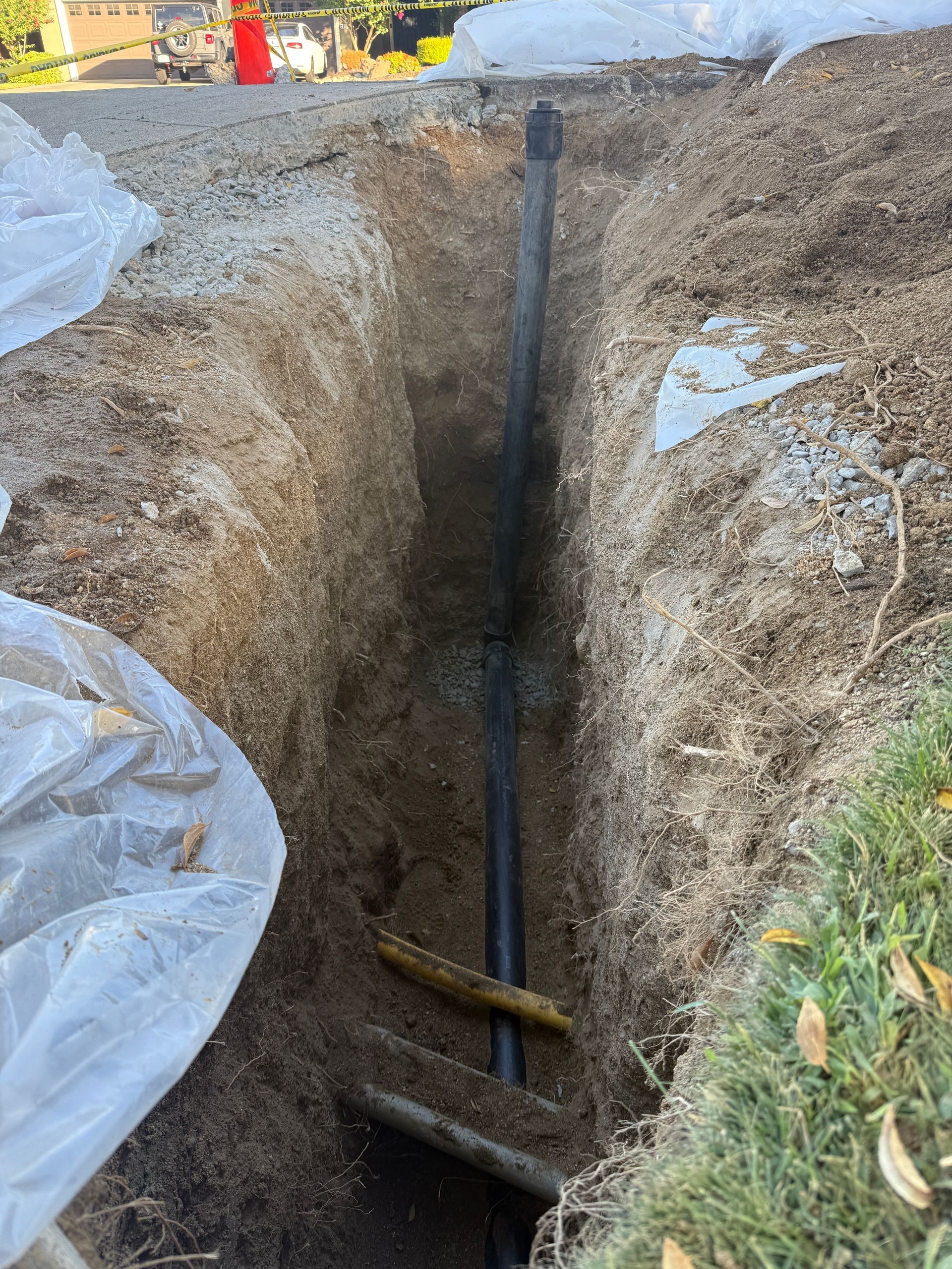 Rightway Plumbing Sewer And Drain - Trench dug in dirt with black pipe running through it; grass and road visible nearby.
