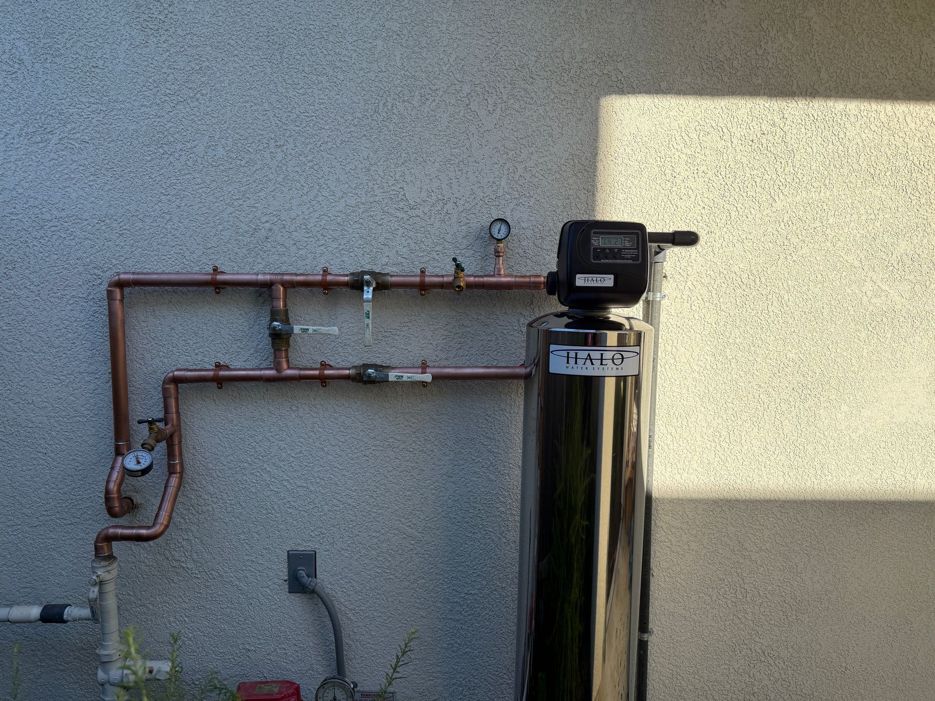 Outdoor water softener with copper pipes mounted on a stucco wall in sunlight