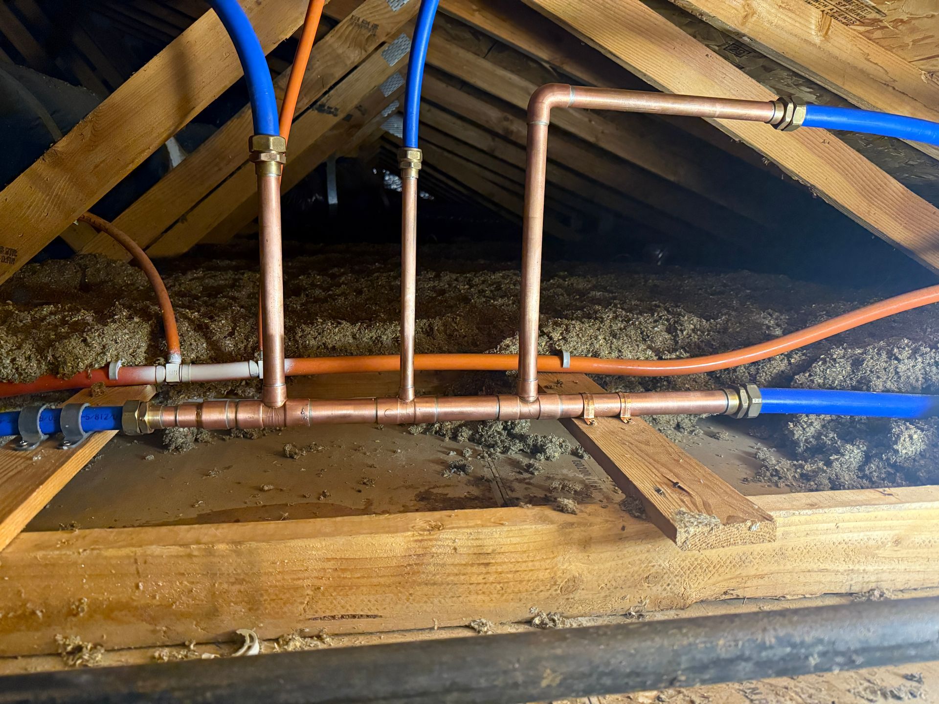 Copper and blue plumbing pipes under a wooden floor in a crawl space
