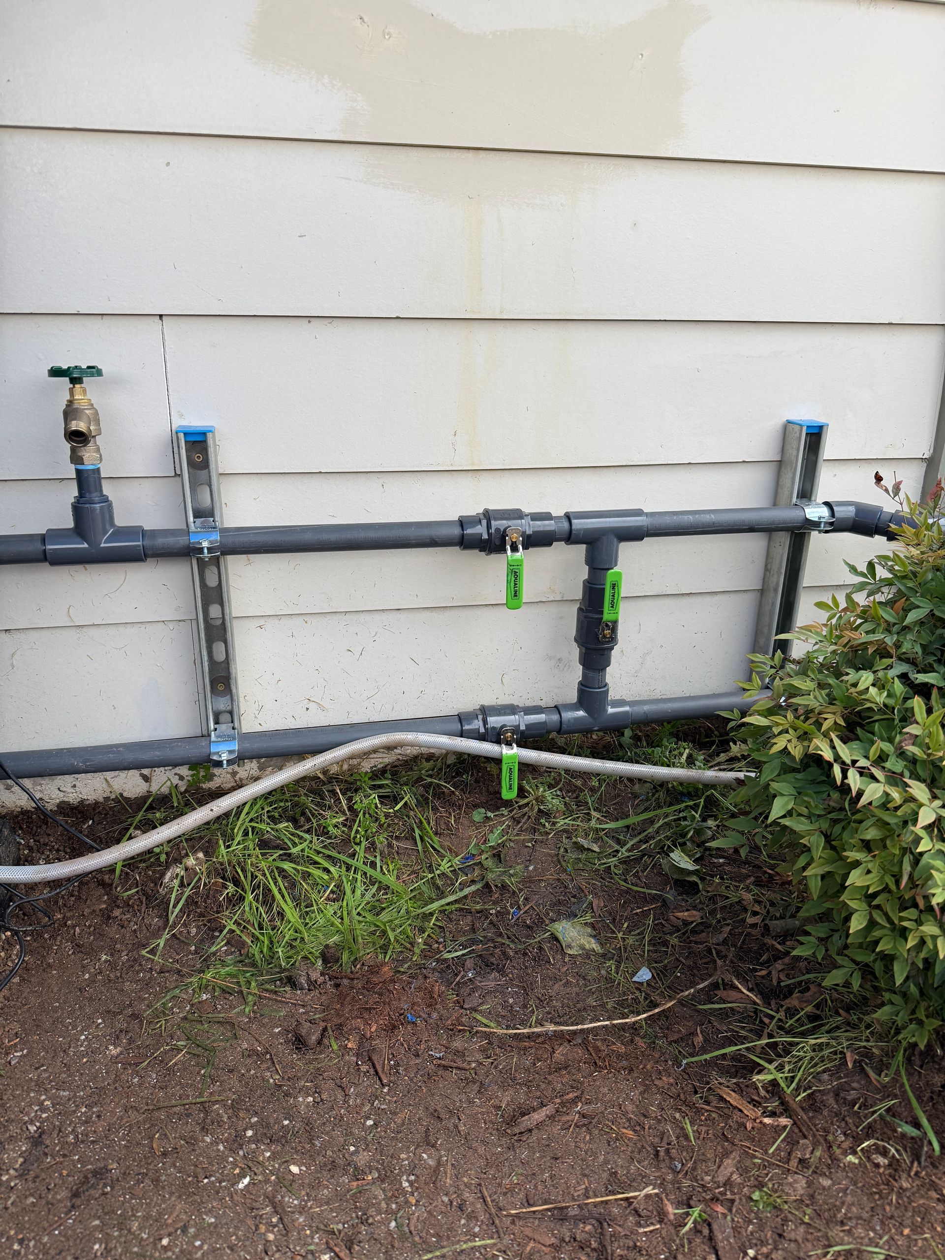 Outdoor plumbing pipes and valves mounted on a beige house wall above dirt and weeds.