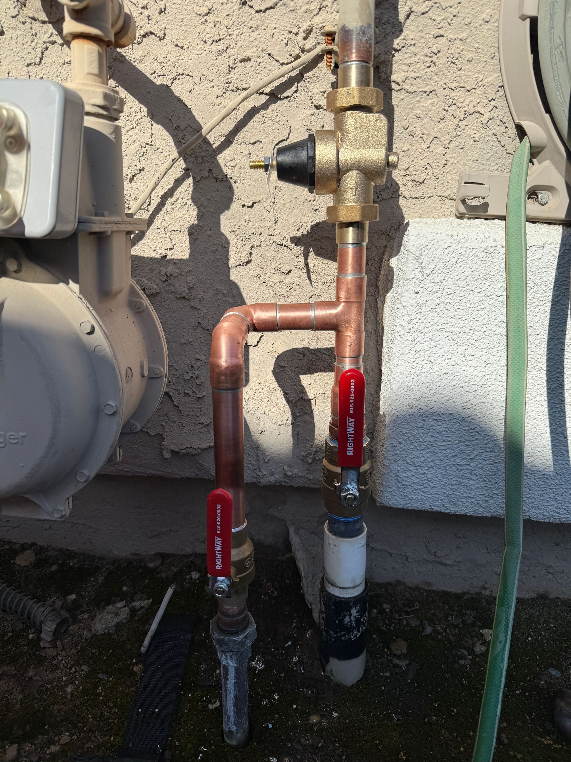 Outdoor plumbing pipes and red-handled valves beside a stucco wall and gas meter