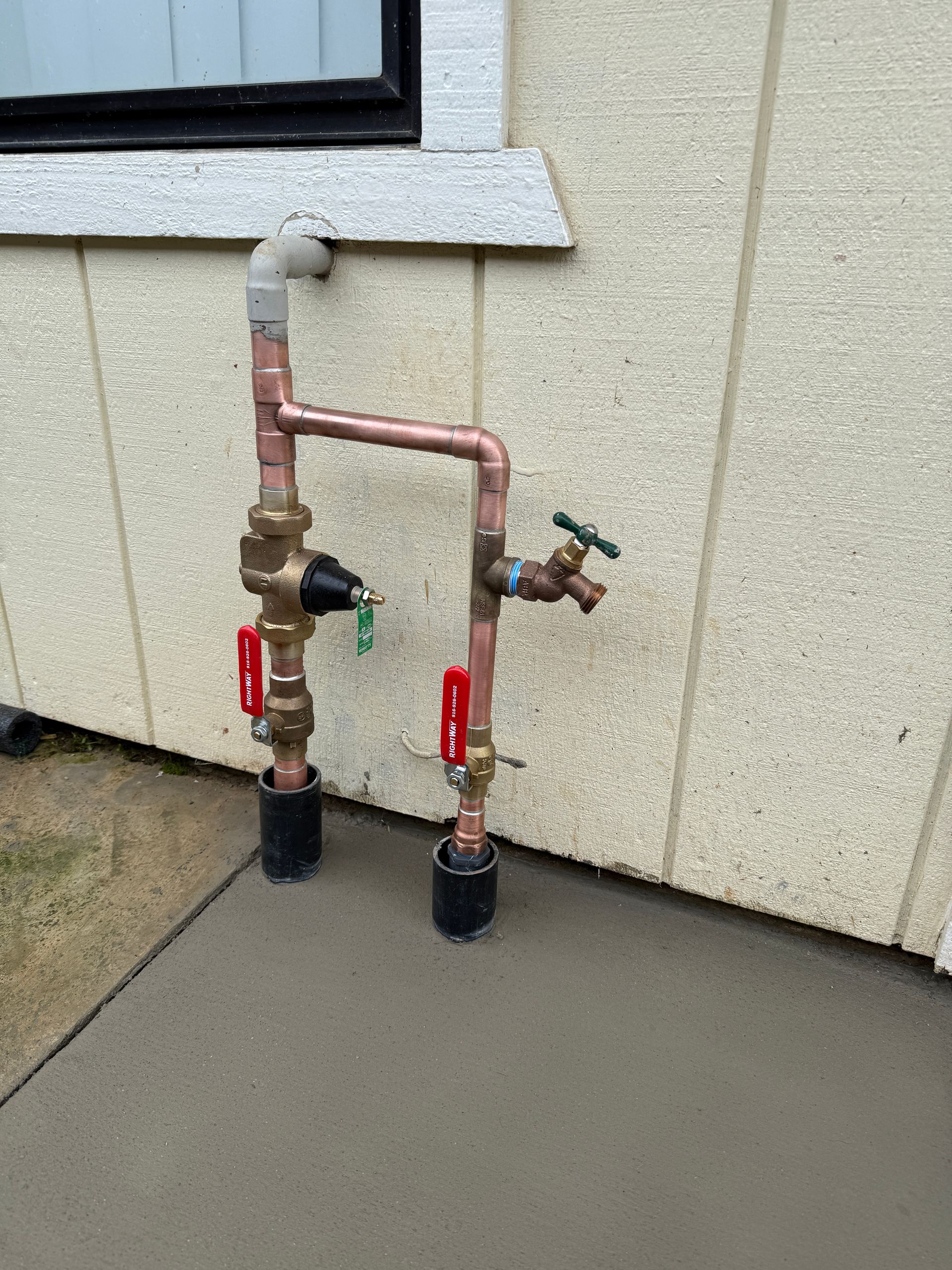 Two outdoor water shutoff valves and pipes mounted on a wall above a concrete slab