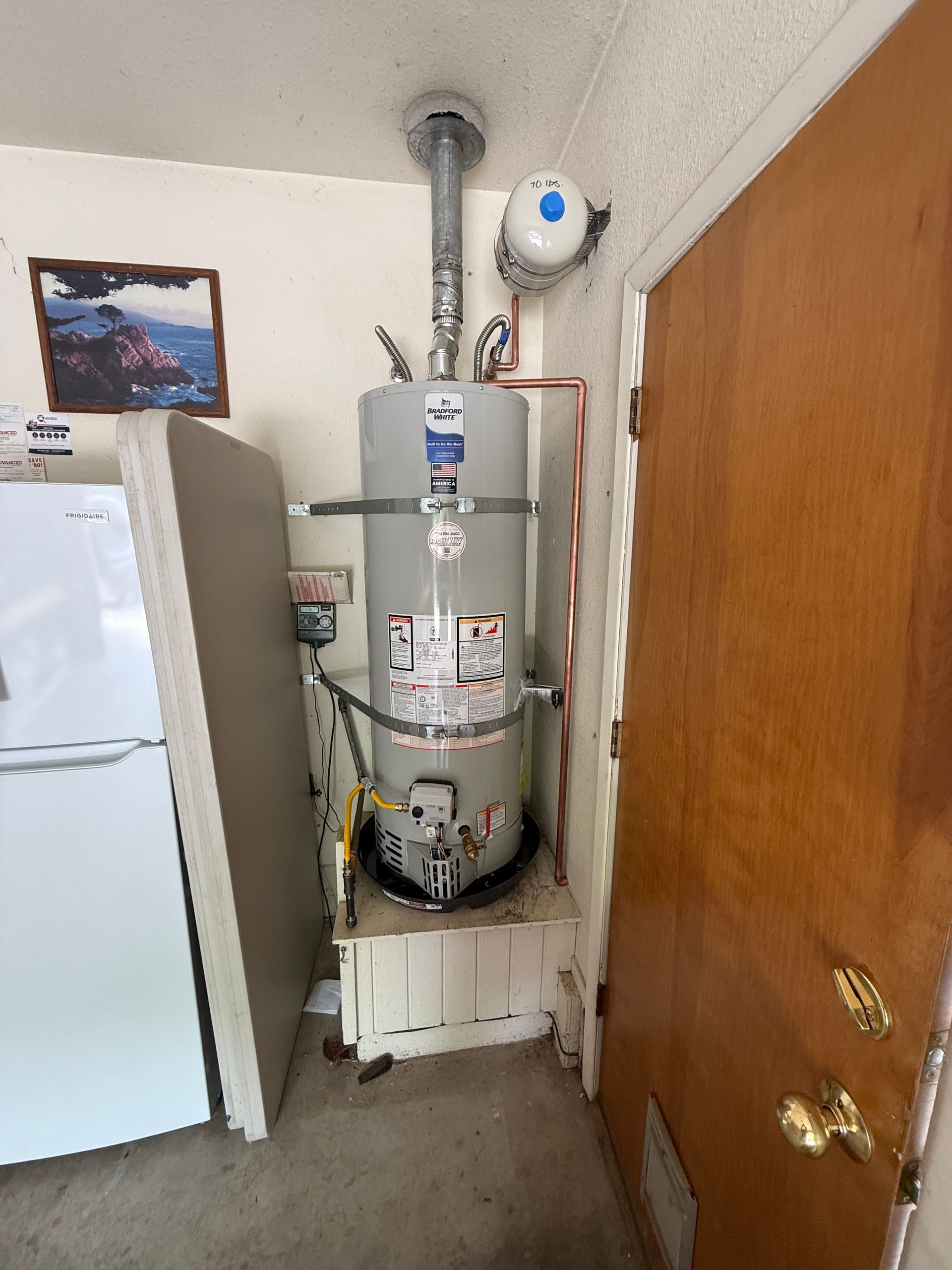 Utility closet with a water heater, refrigerator, and wooden door in a cramped indoor space