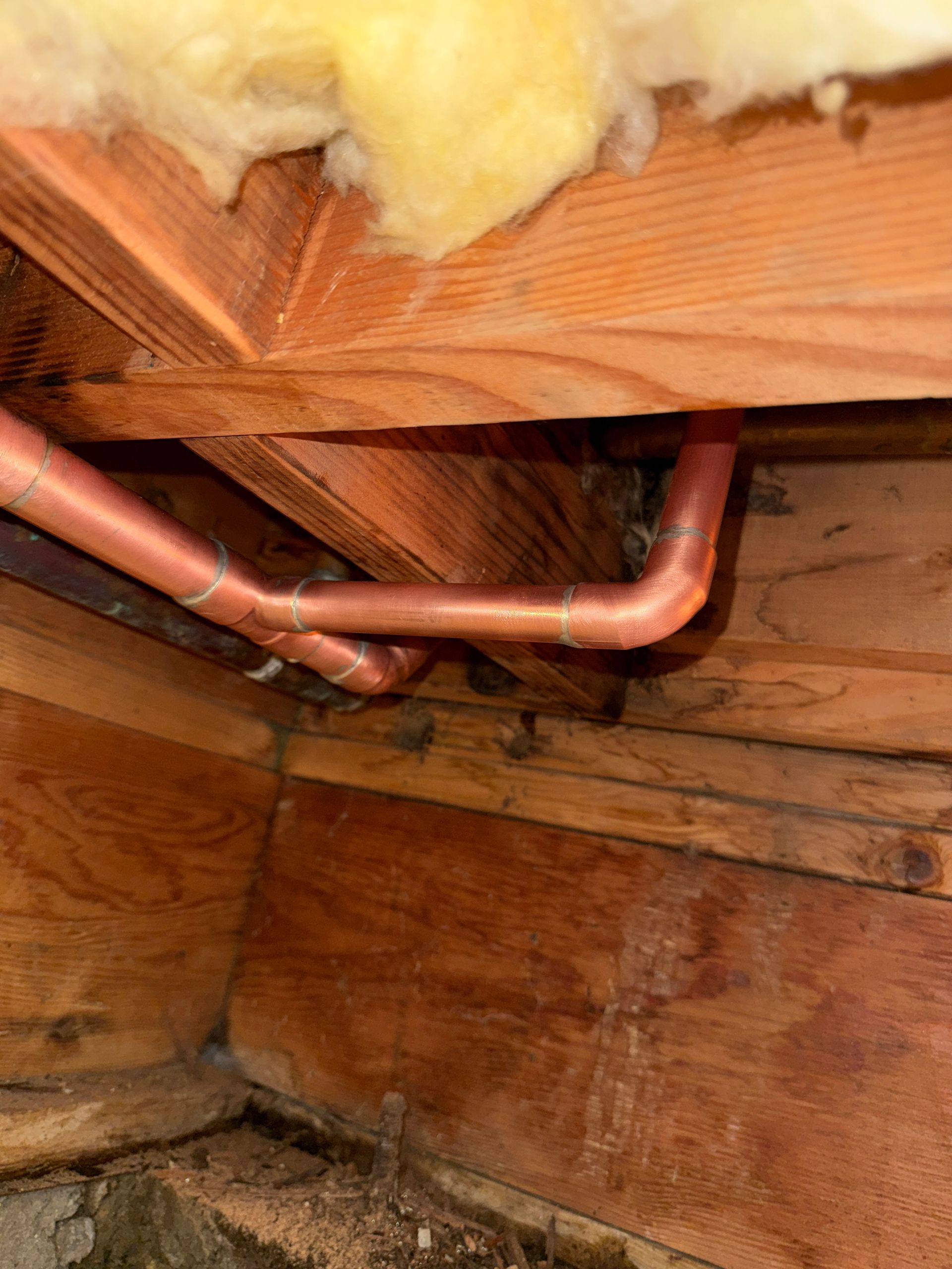 Copper pipes running under wooden floor joists with yellow insulation above.