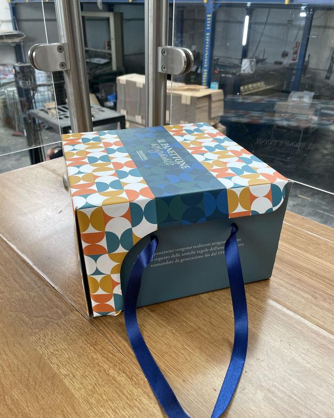 Scatola regalo decorata con manici blu, appoggiata su un tavolo di legno all'interno.