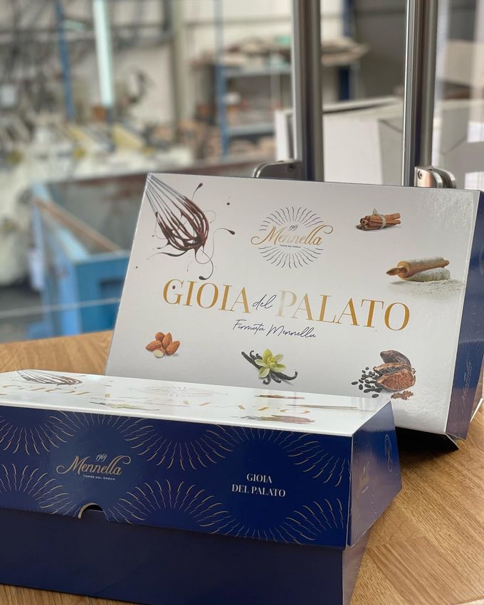 In un negozio, su un tavolo di legno, si vedono due scatole regalo di cioccolatini: una bianca e una blu scuro con scritte dorate.