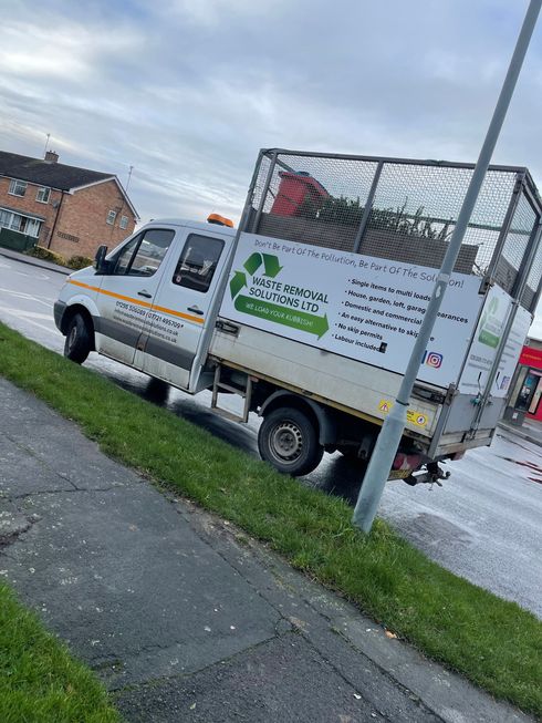 White recycling truck on a roadside, grass verge, with a green logo and cage bed filled.