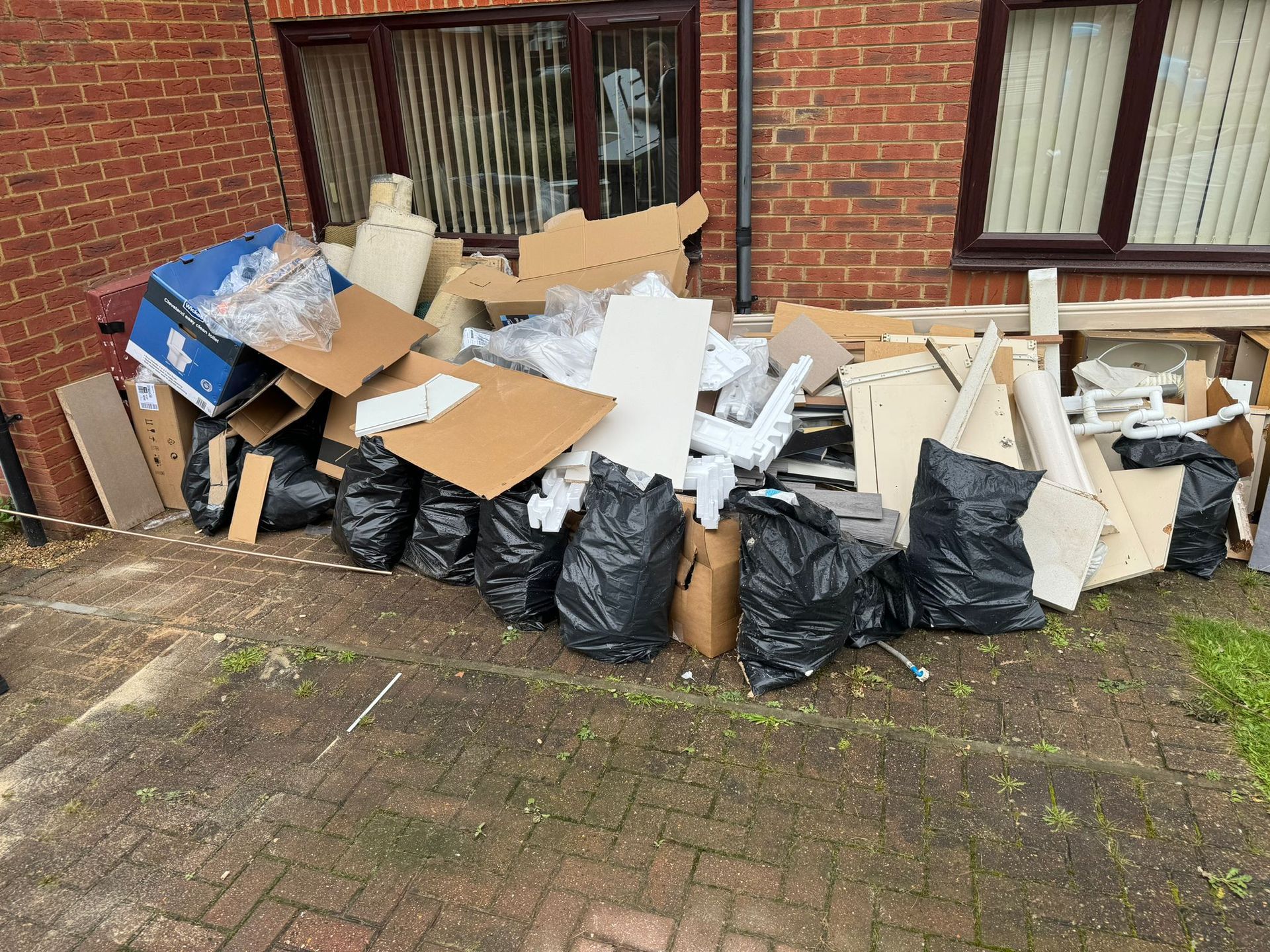 Pile of discarded cardboard boxes, construction debris, and black trash bags on concrete. Brick building background.