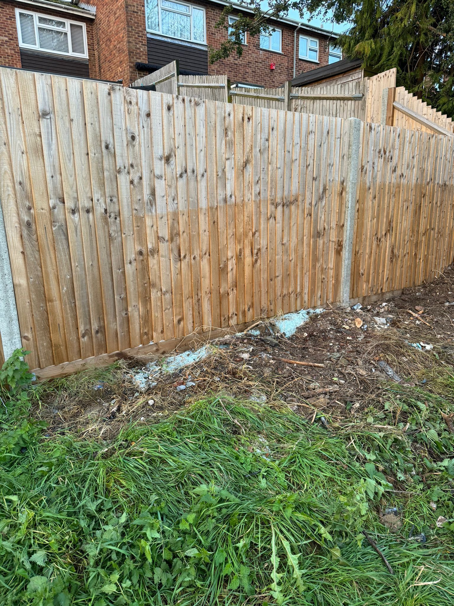 Wooden fence bordering a yard with overgrown weeds in front and a brick building in the background.