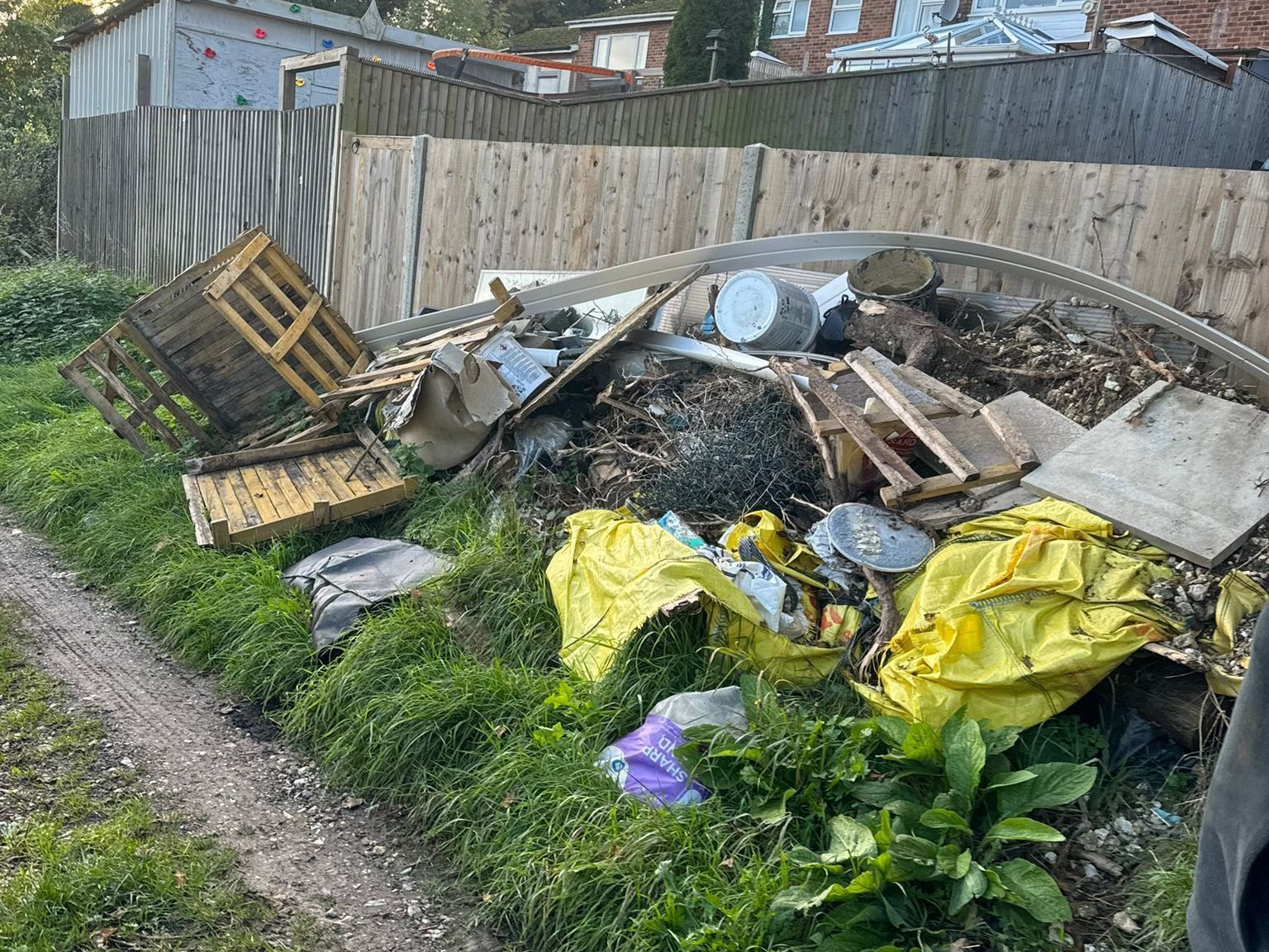 Pile of trash including pallets, metal, and yellow bags along a grassy edge next to a wooden fence.