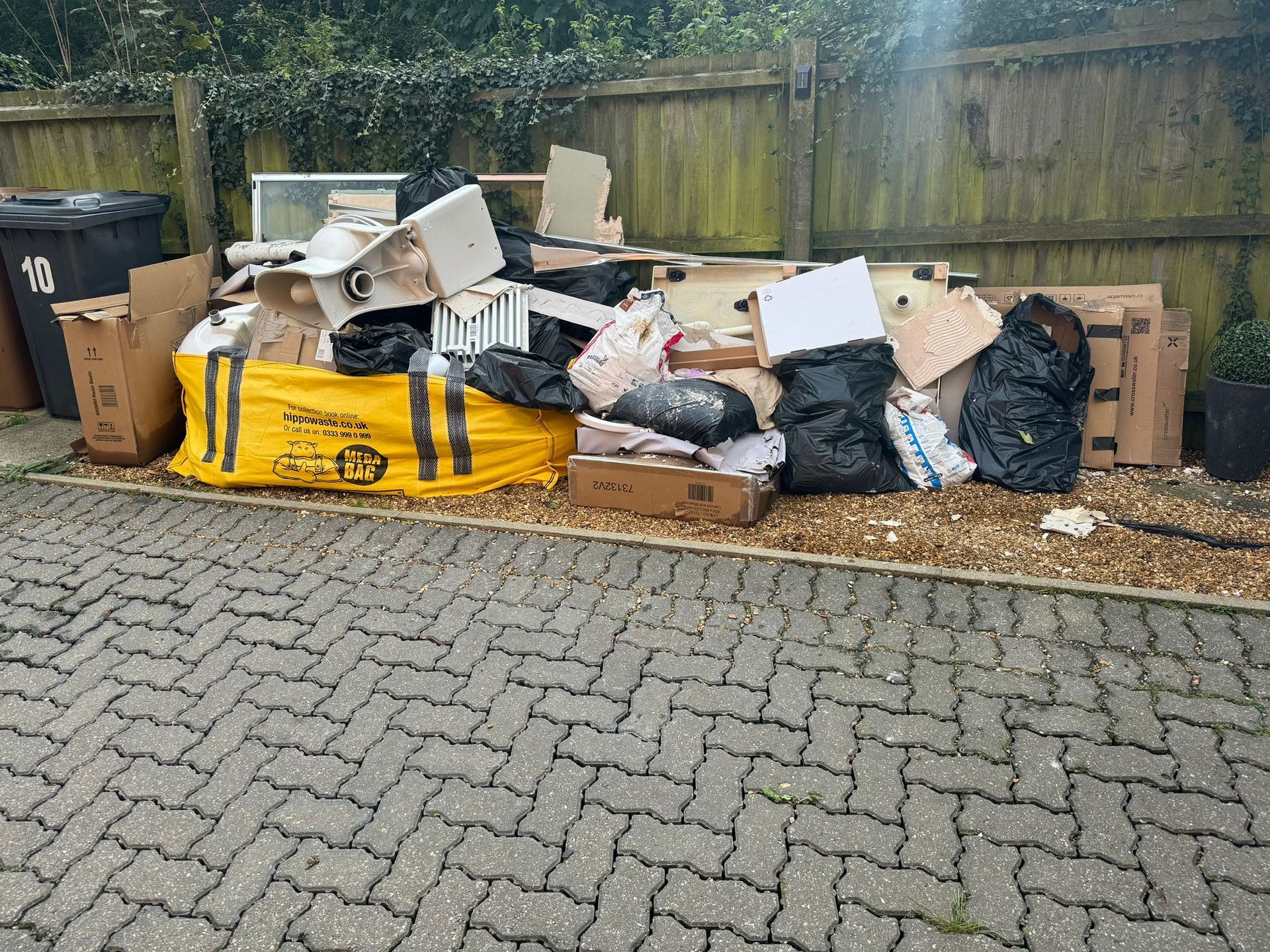 Pile of trash, including boxes, black bags, and building materials, on gravel by a fence.