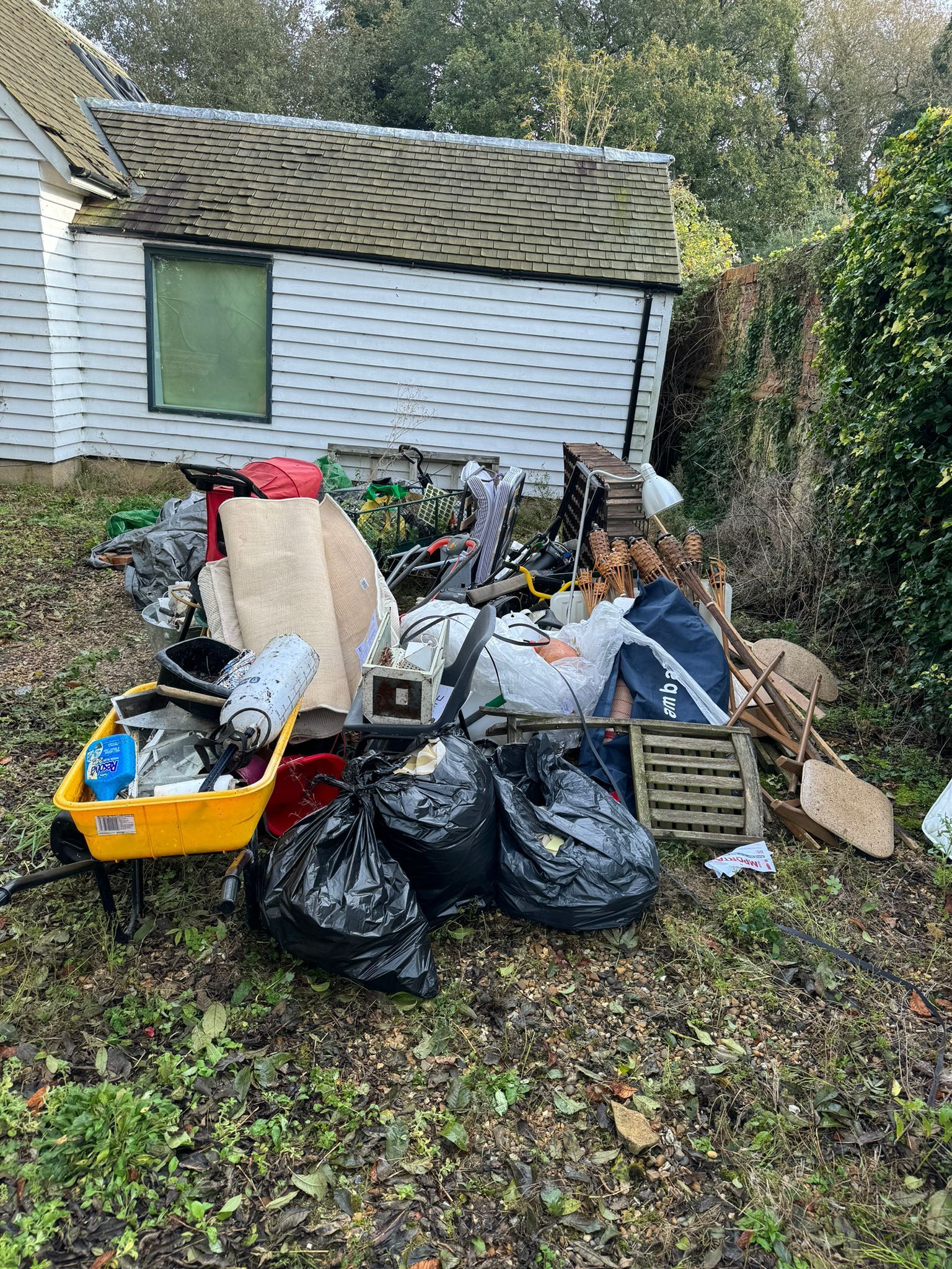 Pile of trash and debris in front of a white shed. Includes bags, wheelbarrow, and furniture. Outdoors.