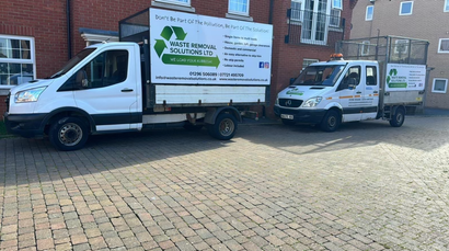 Two white recycling trucks parked on a brick road with a building in the background.