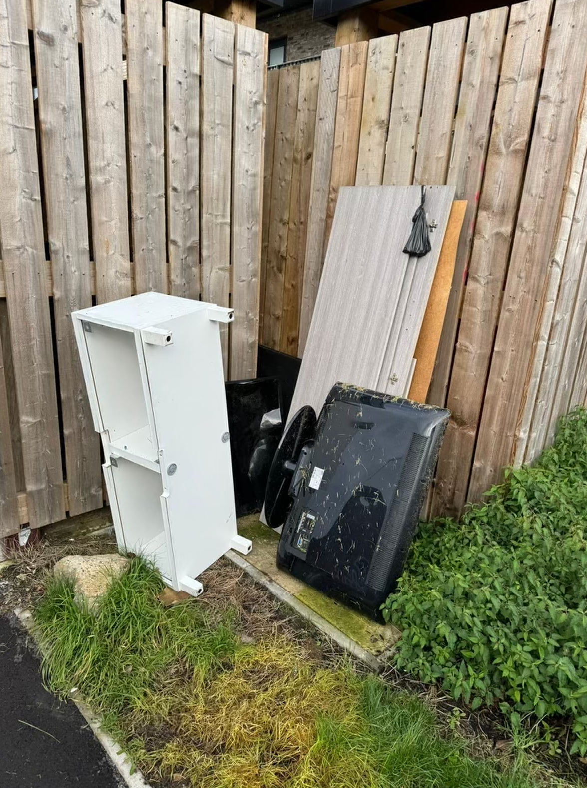 White shelving unit, discarded electronics and wood debris next to a wooden fence.