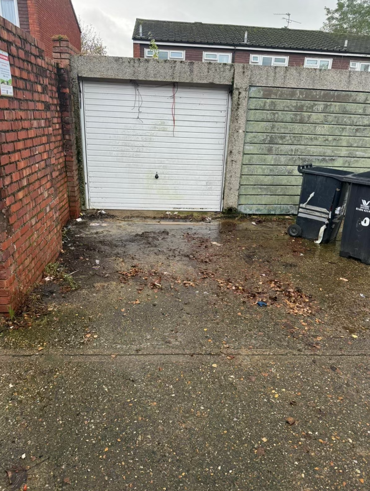 Exterior view of a weathered garage with a white door, next to a brick wall and a green concrete wall, with trash cans in the foreground.