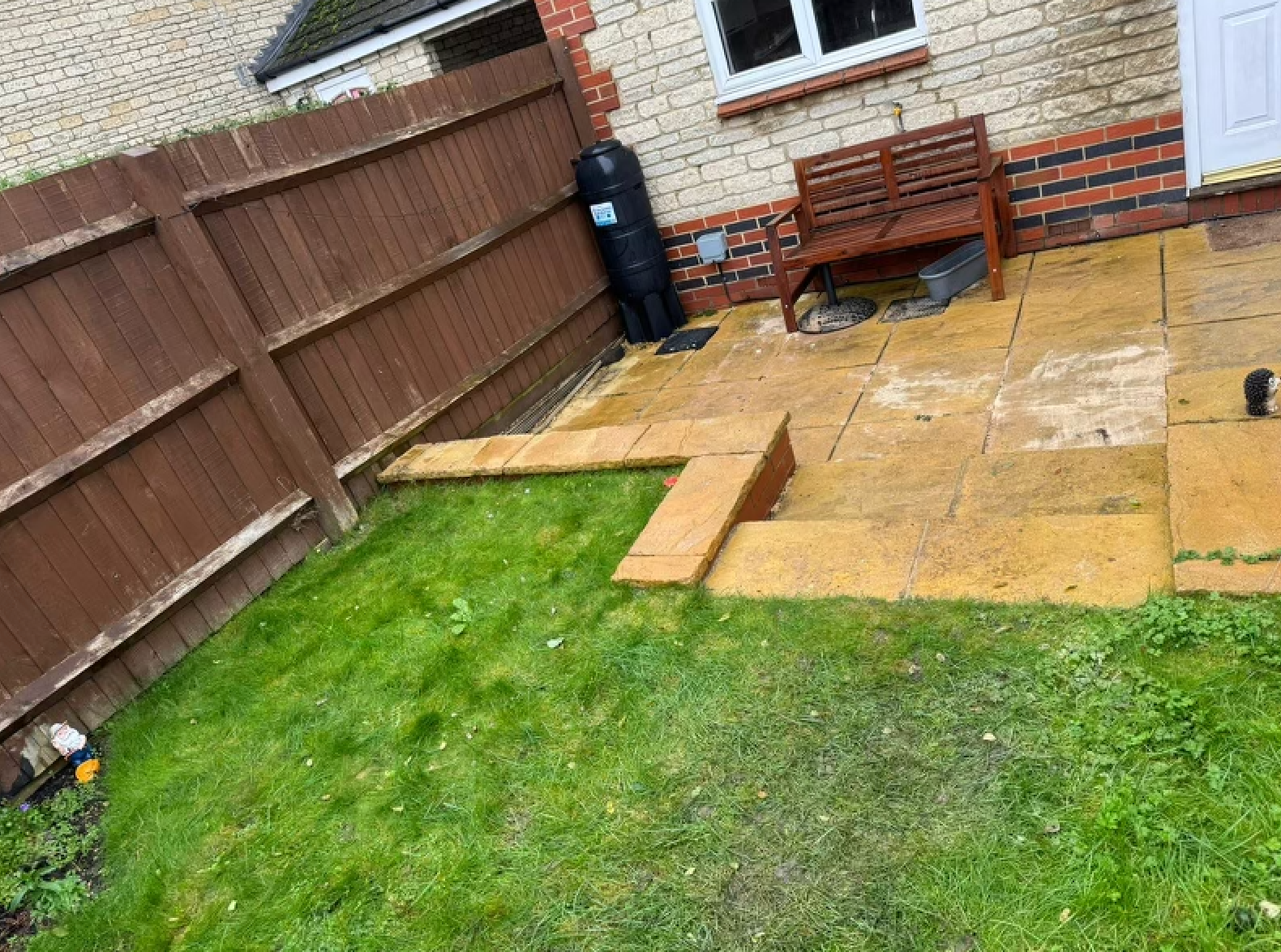 Backyard patio with yellow stone pavers, brown fence, and a wooden bench. Green grass surrounds.