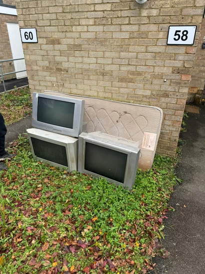 Three old TVs and a mattress lean against a brick wall beneath house number signs.