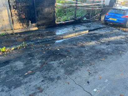 Charred debris and water on asphalt near burnt structure and blue car.