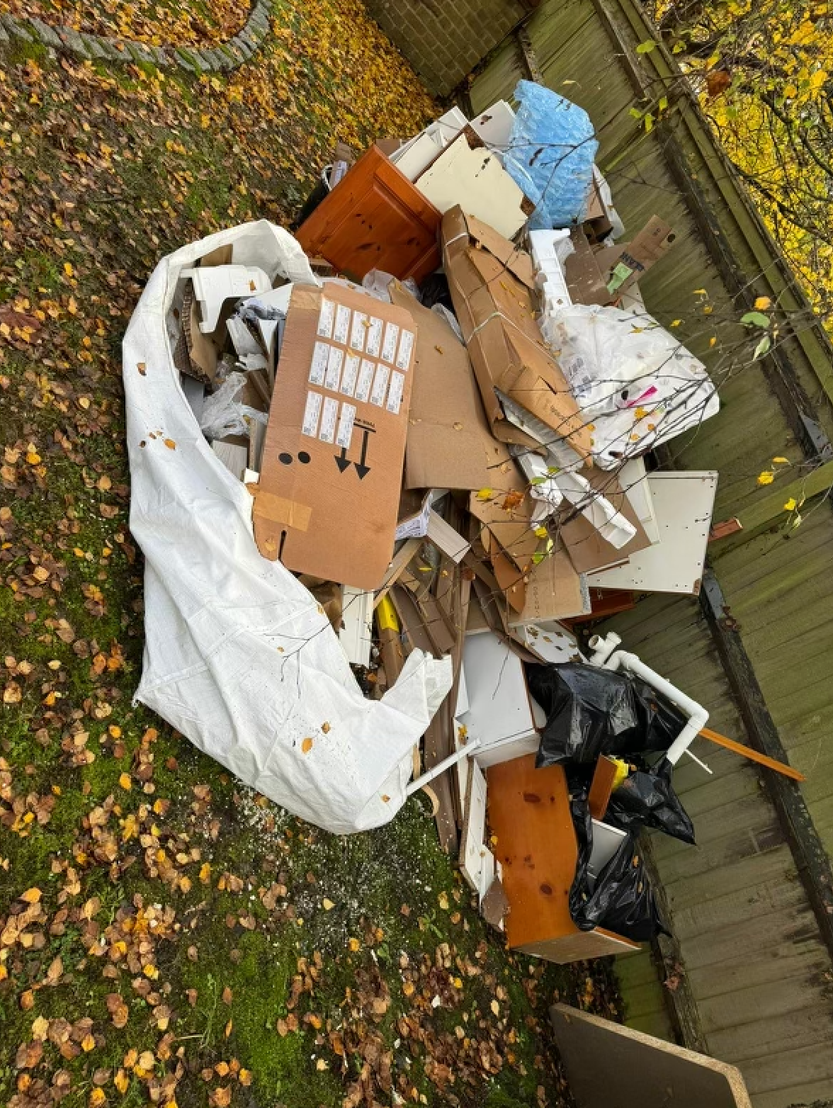 Pile of assorted trash in a yard, including cardboard, wood, and plastic bags; autumn leaves and wooden fence.