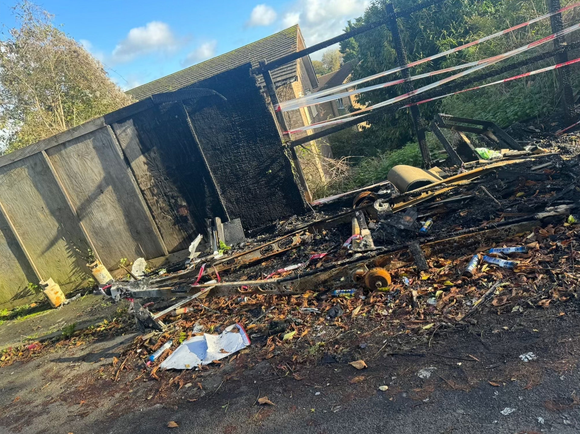 Burnt shed next to fence. Debris and charred wood cover ground. White and red tape strung nearby.