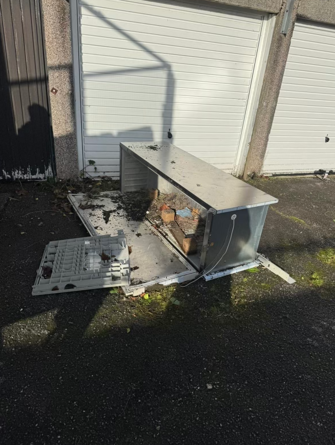 A damaged metal box lying open on asphalt in front of garage doors. Debris surrounds it.