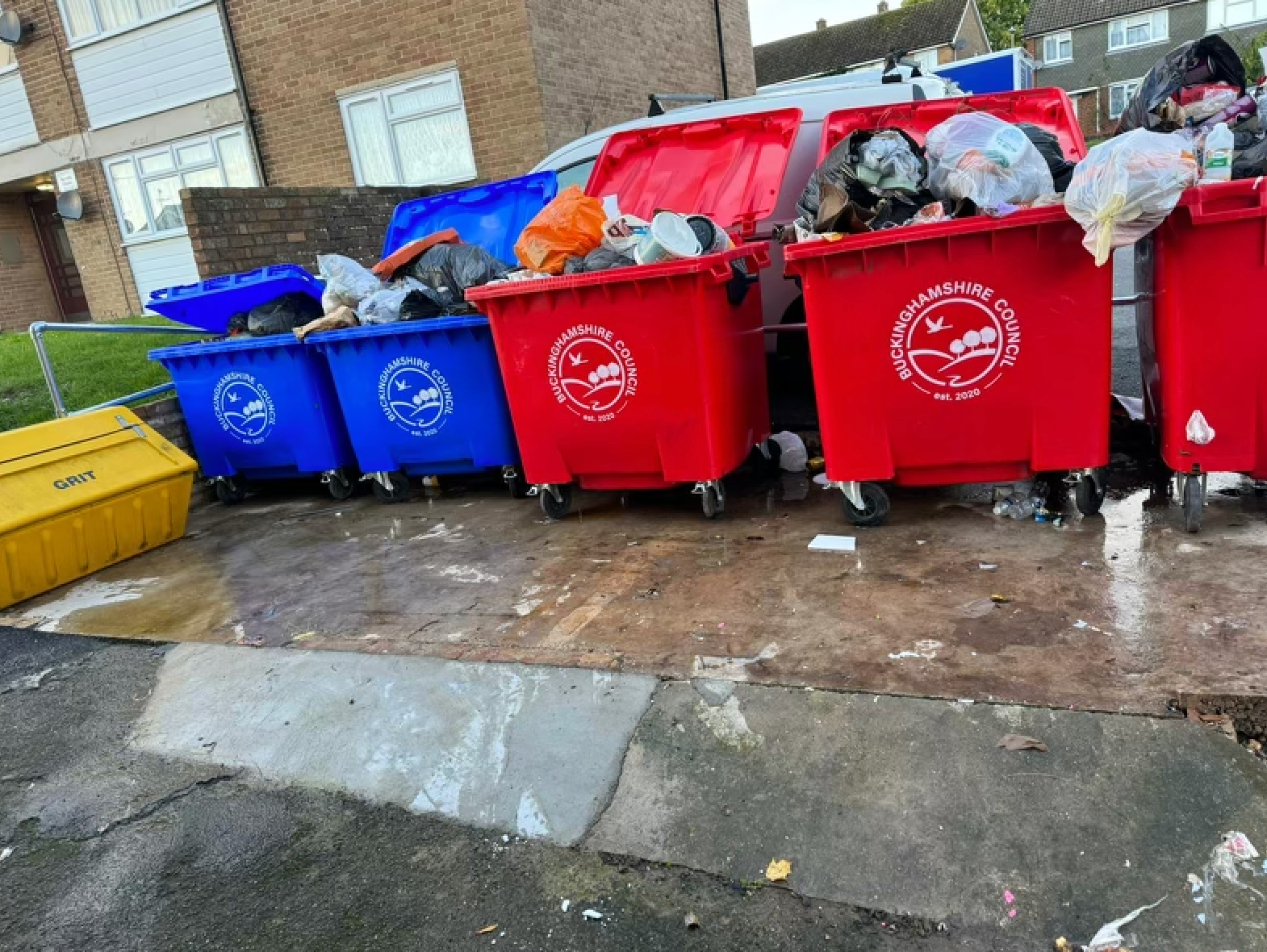 Overflowing blue and red trash bins with scattered refuse on a sidewalk. A yellow bin is on the left.