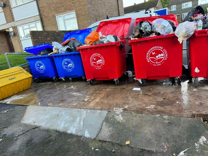 Overflowing red and blue bins; trash scattered around; yellow bin on the left; residential setting.