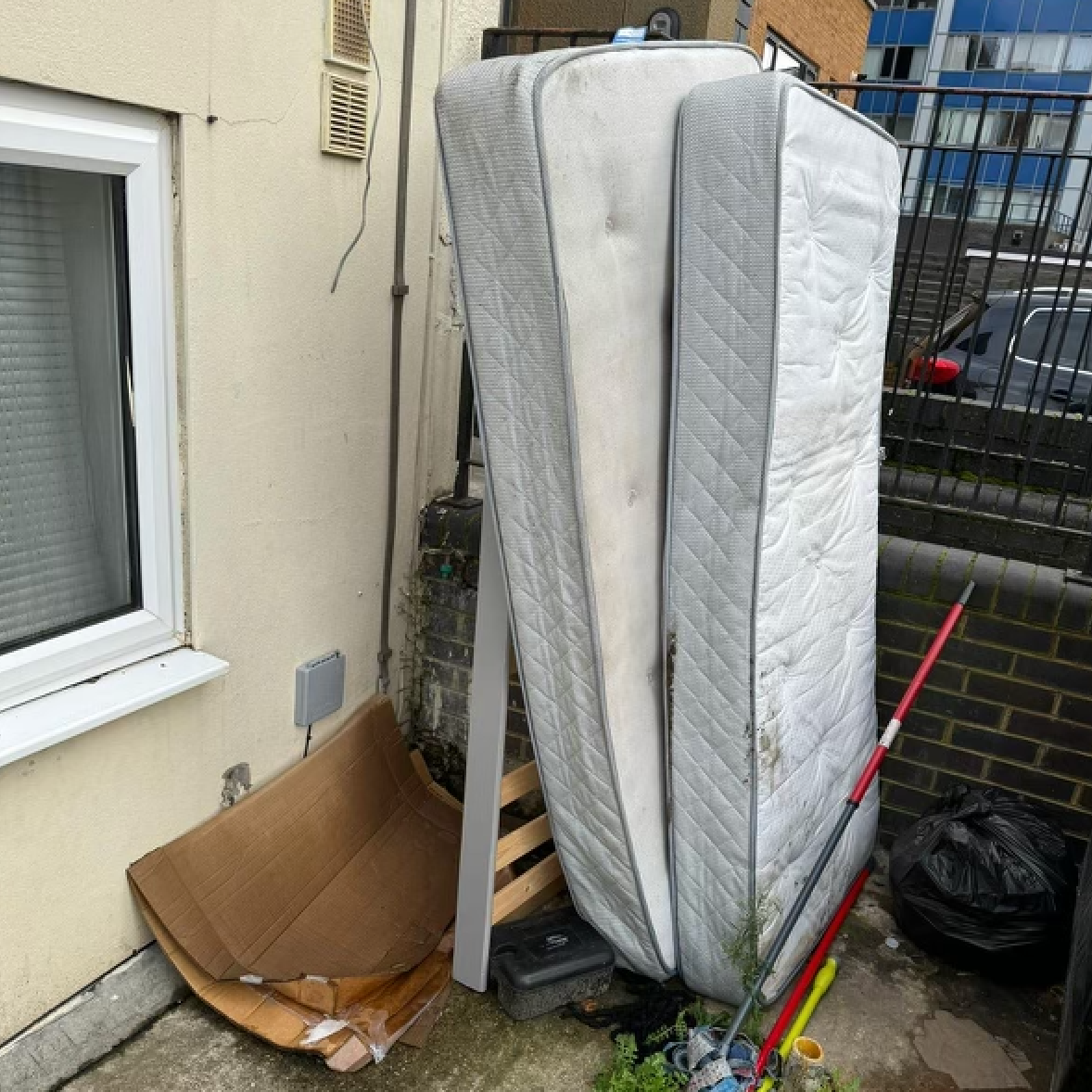 Mattress leaning against a wall, next to trash and cardboard. Outdoor setting.