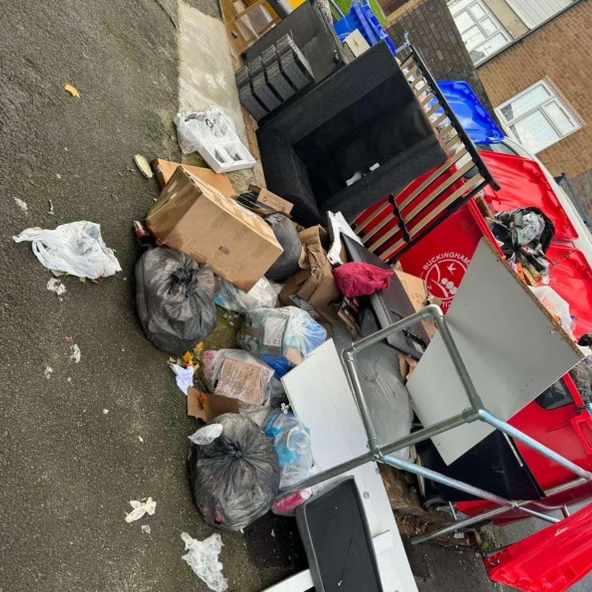 Pile of household waste including bags, boxes, and furniture on a paved area, likely outdoors.