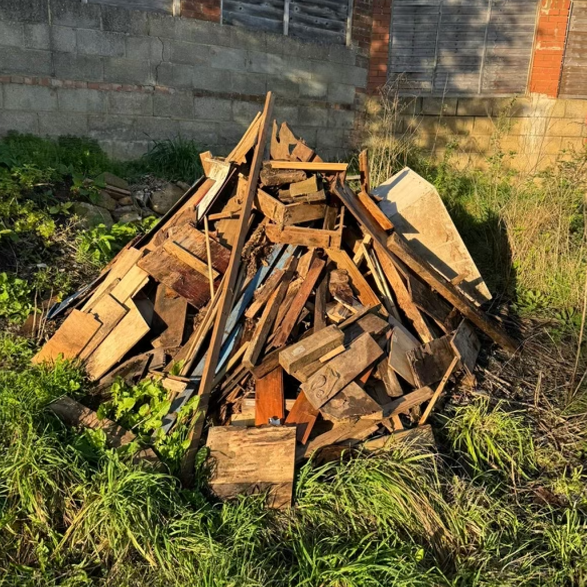 Pile of weathered wood scraps in tall grass, against a concrete and brick wall.