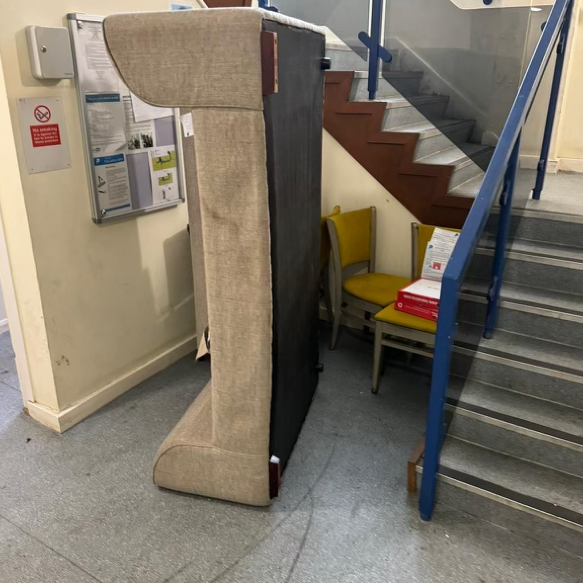 Couch blocking a staircase in a building. Yellow chair and paperwork are behind it. Beige walls, stairs, and gray floor.