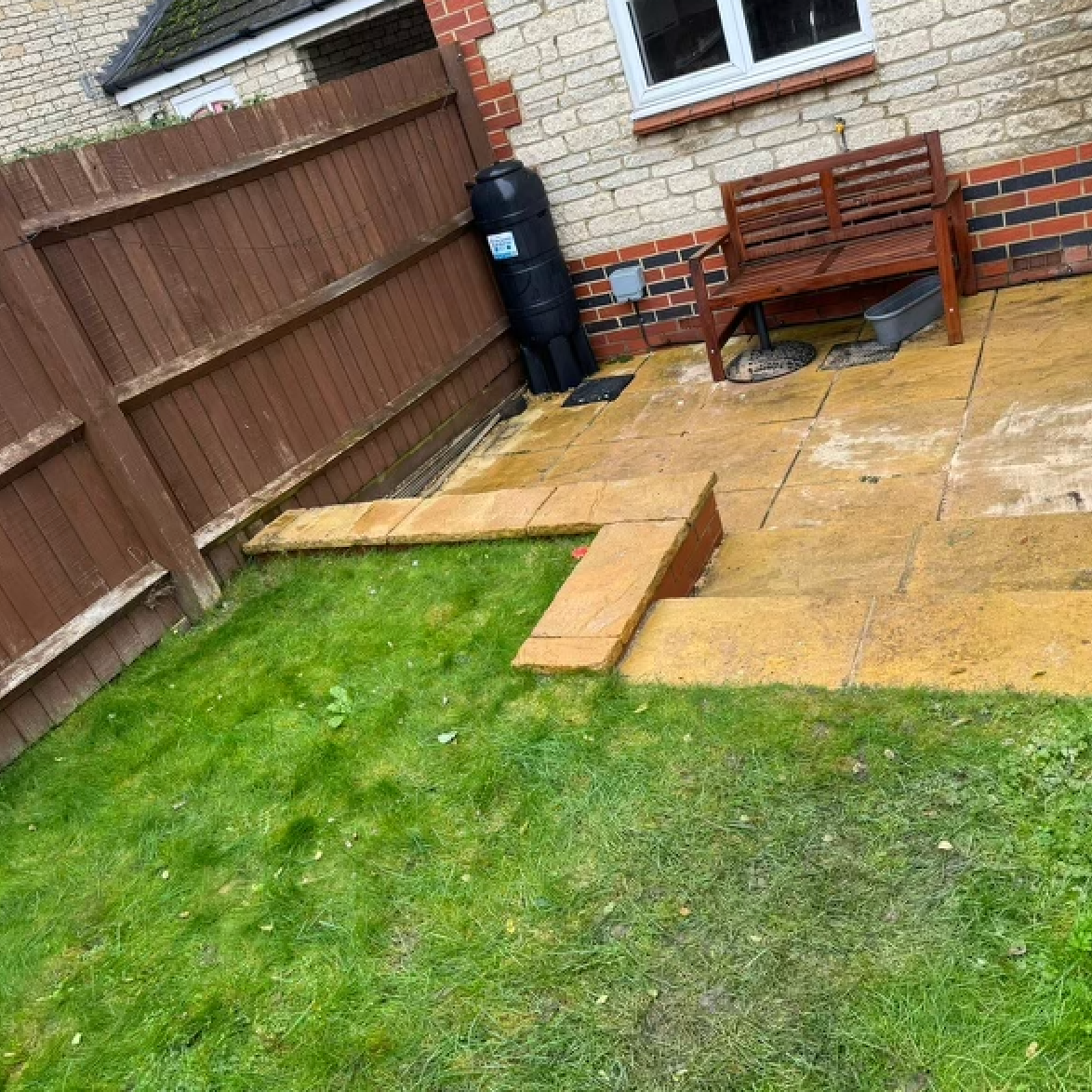 A backyard with a patio, lawn, and wooden fence. There's a bench, water barrel, and paving stones.