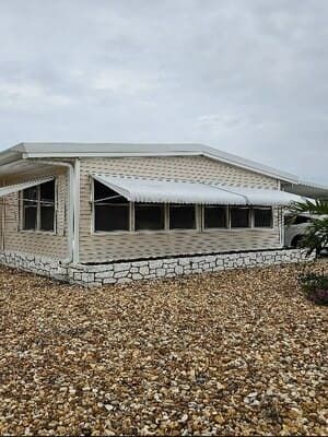 White house with stone foundation and covered porch, set on a rocky lot under a cloudy sky