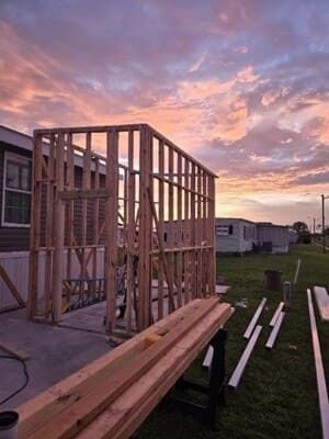 Wooden house framing at sunset with lumber stacked in the foreground and a grassy yard behind.