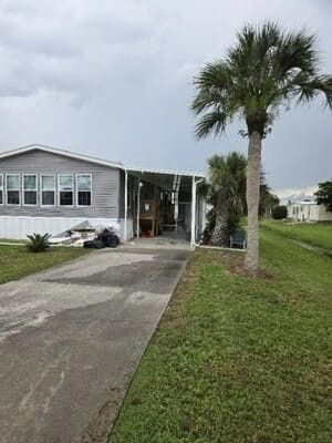 Single-story house with driveway, carport, and palm tree under cloudy sky.