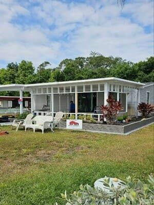 White mobile home with screened porch, lawn chairs, and flowers on a grassy lot under a cloudy sky
