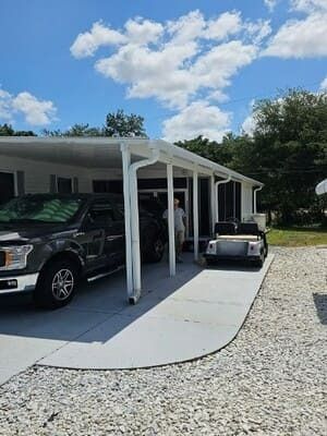 Covered driveway with parked vehicles beside a white house on a sunny day