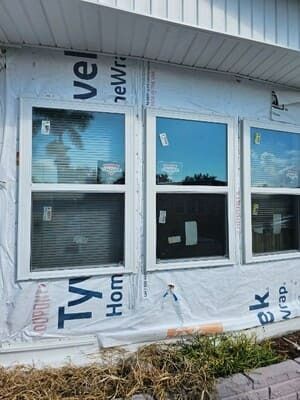 House exterior under construction with three windows, wrapped in white Tyvek siding and blue trim.