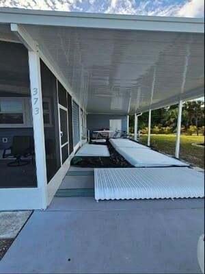 Covered patio with white roll-down shades, a table, chairs, and a concrete walkway beside a house