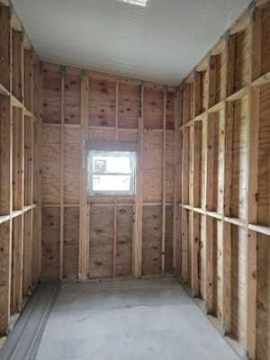 Unfinished narrow room with exposed wood framing, white walls, and a small window at the far end.