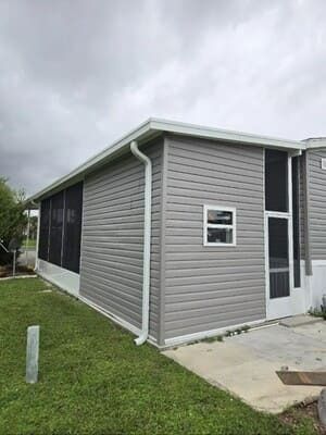 Gray single-story house with white trim, screened windows, and a small concrete patio beside a grassy yard