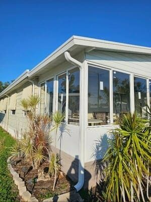 White sunroom addition with large windows and potted plants beside a house under a blue sky