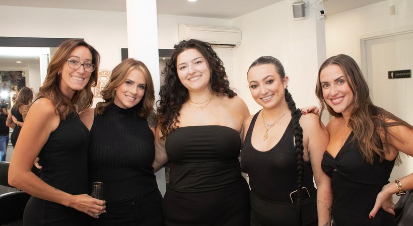 Five women in black dresses smiling and posing together at an indoor event.