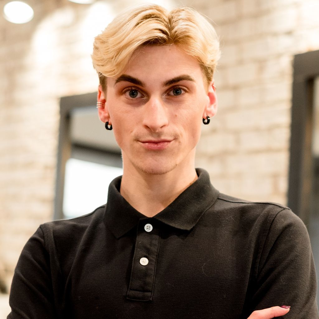 Person with blonde hair and black polo shirt, arms crossed, standing in a bright indoor hallway.