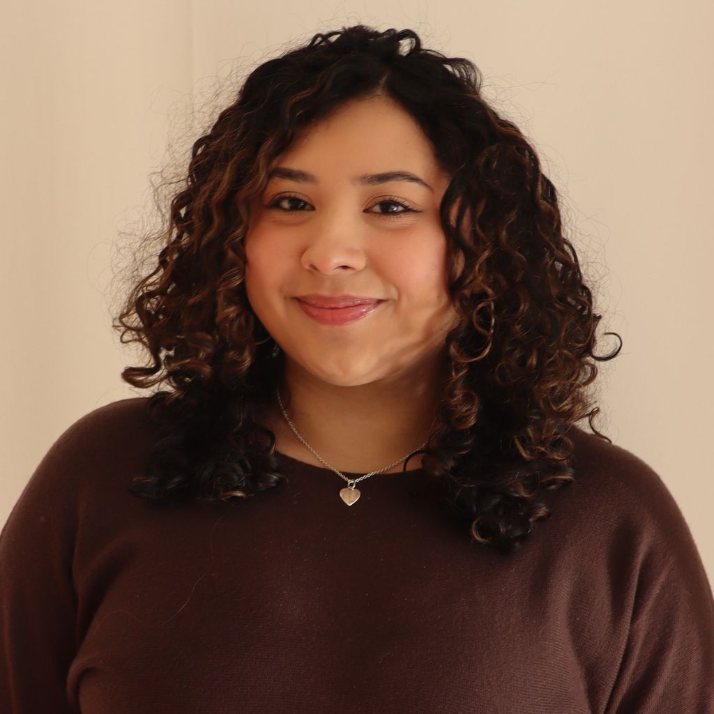 Person with curly hair smiling in a brown sweater against a beige background