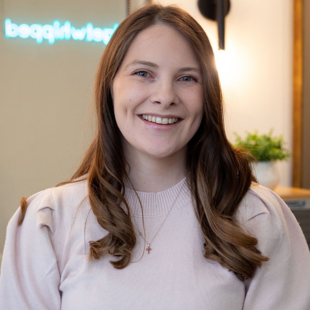 Smiling woman in a light pink sweater standing indoors, with a blurred office background and plants.