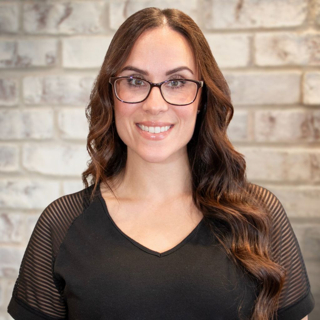 Smiling person with dark hair and red streaks, wearing a black top against a stone wall.